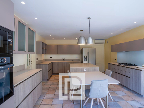 Modern open kitchen with grey cabinetry, a central island, and a dining table with light gray chairs; stainless steel fridge and built-in ovens.