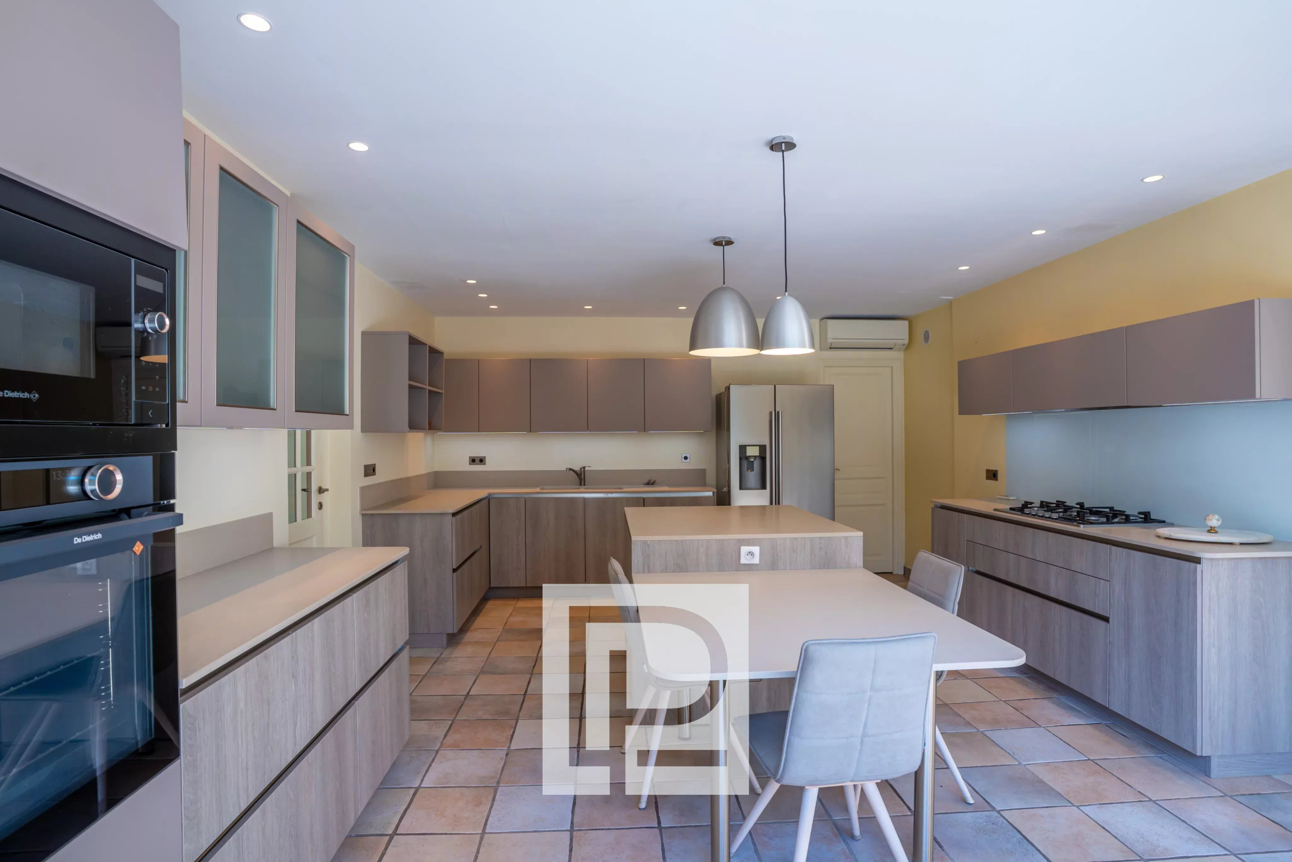 Modern open kitchen with grey cabinetry, a central island, and a dining table with light gray chairs; stainless steel fridge and built-in ovens.