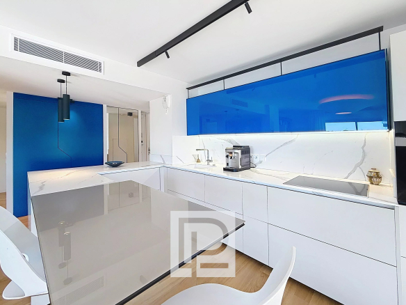 Modern white kitchen with marble countertops, blue glass backsplash, and a glass-topped island.