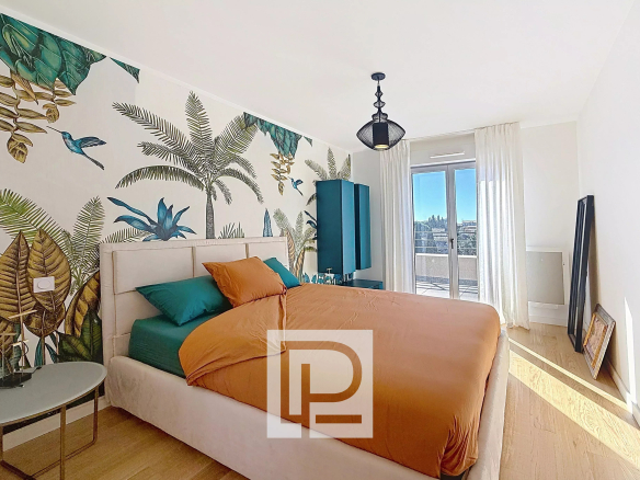 Bright modern bedroom with a botanical mural behind a beige upholstered bed, orange duvet and teal pillows, and a teal wardrobe by the balcony doors.