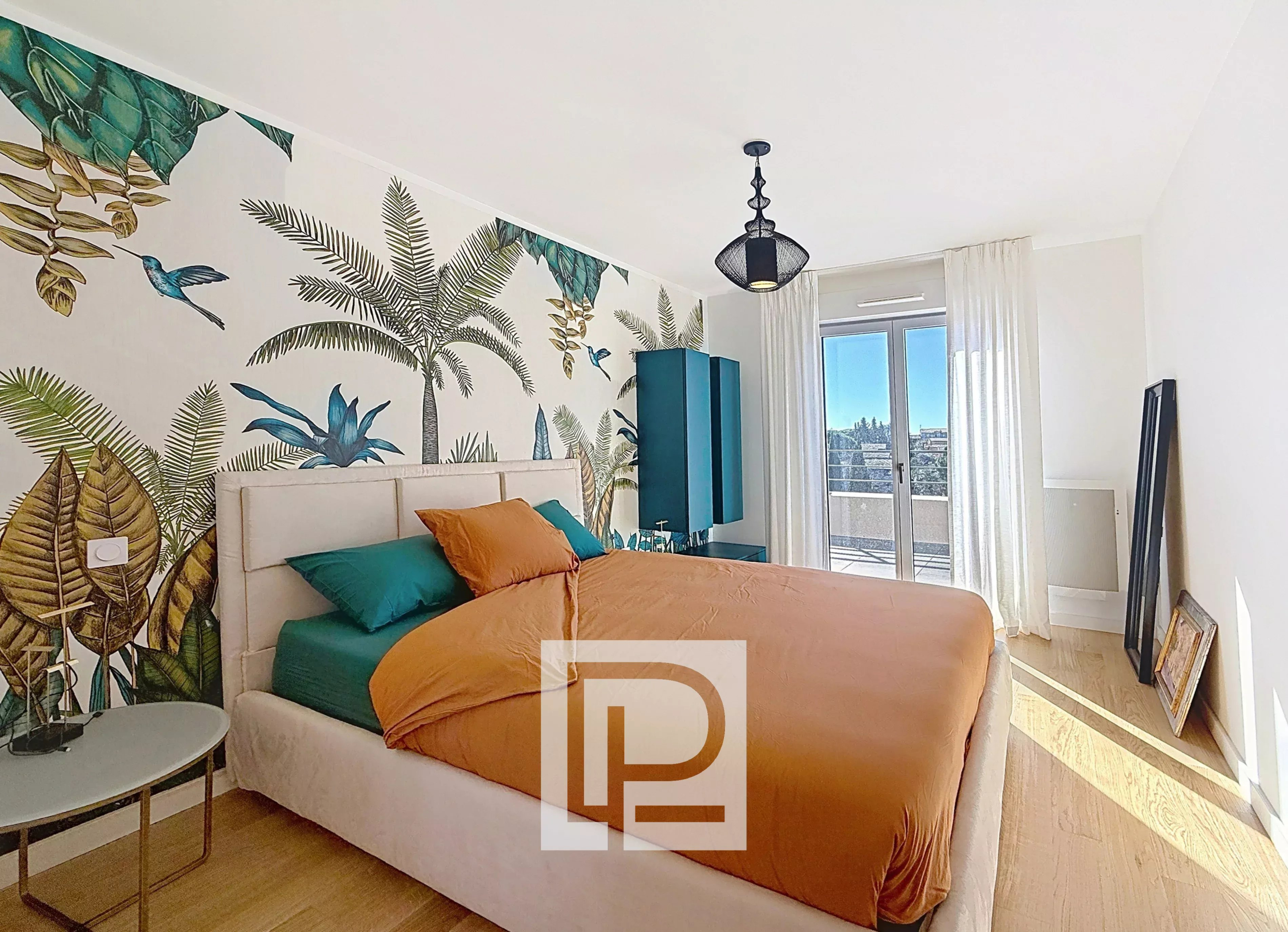 Bright modern bedroom with a botanical mural behind a beige upholstered bed, orange duvet and teal pillows, and a teal wardrobe by the balcony doors.