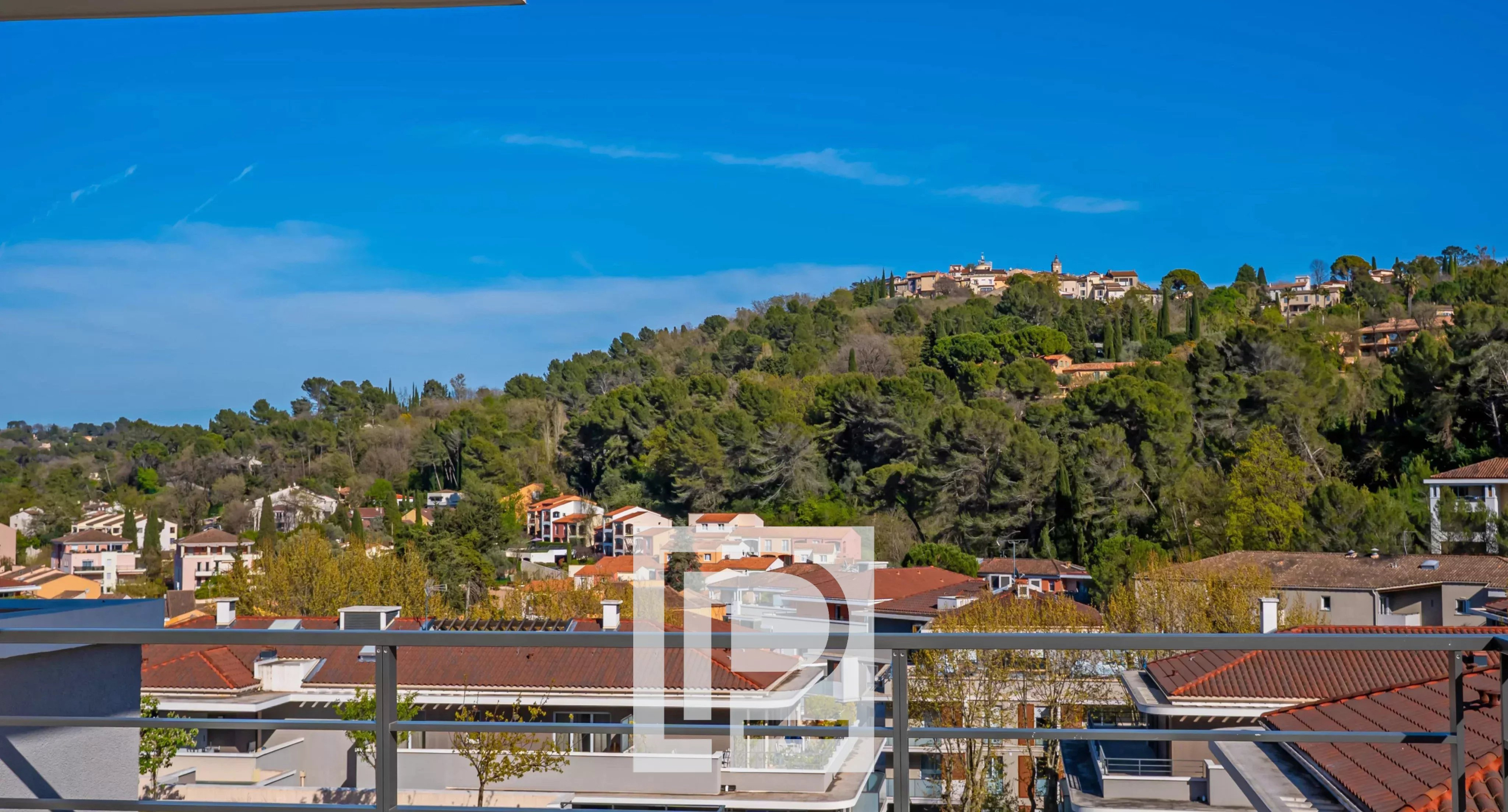 View from a balcony over rooftops toward a tree-covered hillside under a clear blue sky; a metal railing is in the foreground.