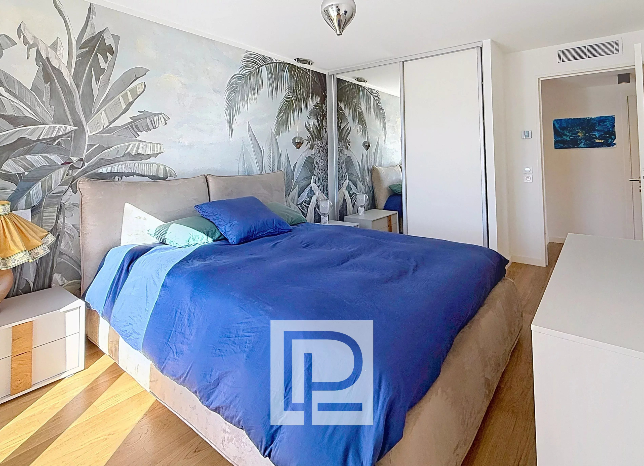 Bedroom featuring a tropical palm mural, a blue duvet and pillows on a beige upholstered bed, white nightstands, and mirrored sliding closet doors.