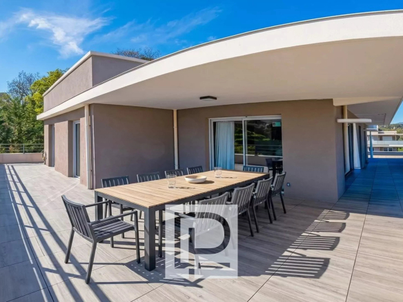 Wide terrace with a long outdoor dining table and black chairs under a curved white overhang, glass doors behind.