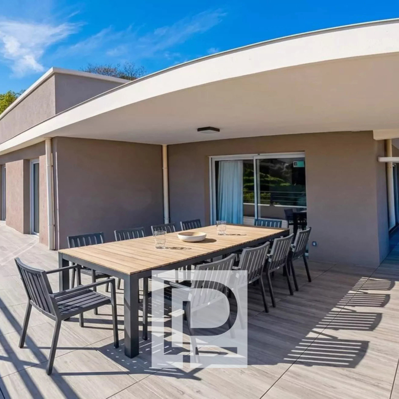 Wide terrace with a long outdoor dining table and black chairs under a curved white overhang, glass doors behind.