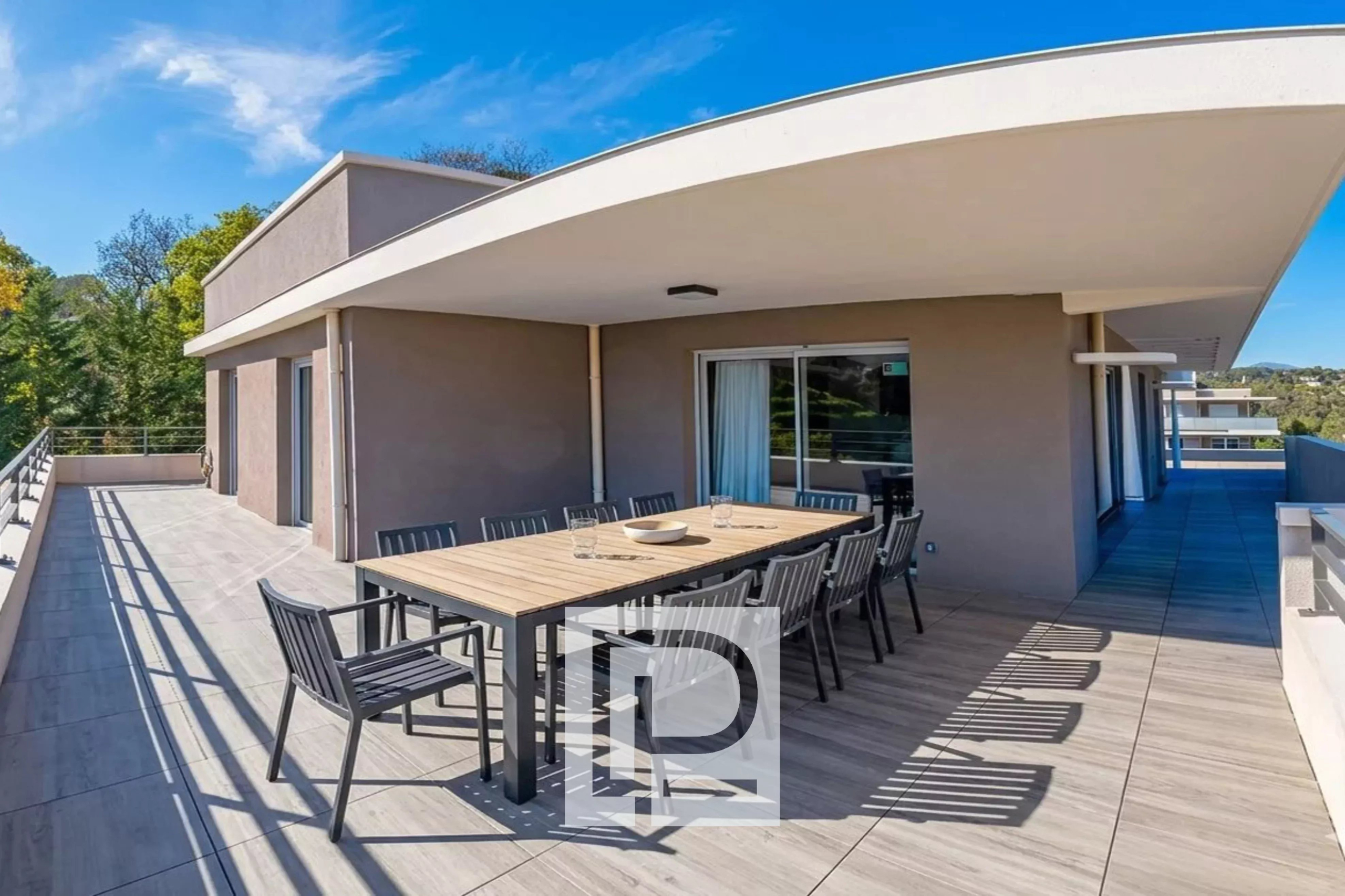 Wide terrace with a long outdoor dining table and black chairs under a curved white overhang, glass doors behind.