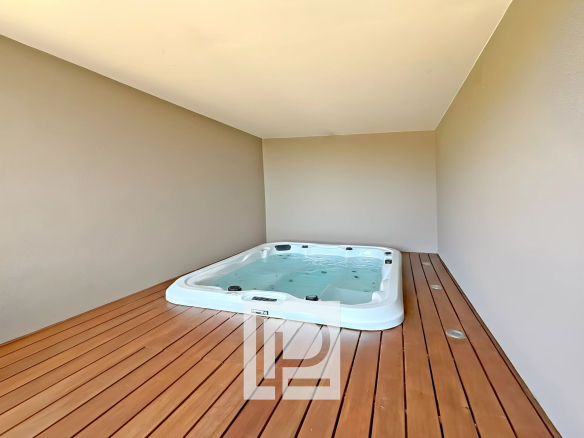 White rectangular hot tub filled with water sits on a dark wood deck inside a beige-walled, minimal outdoor patio or balcony.