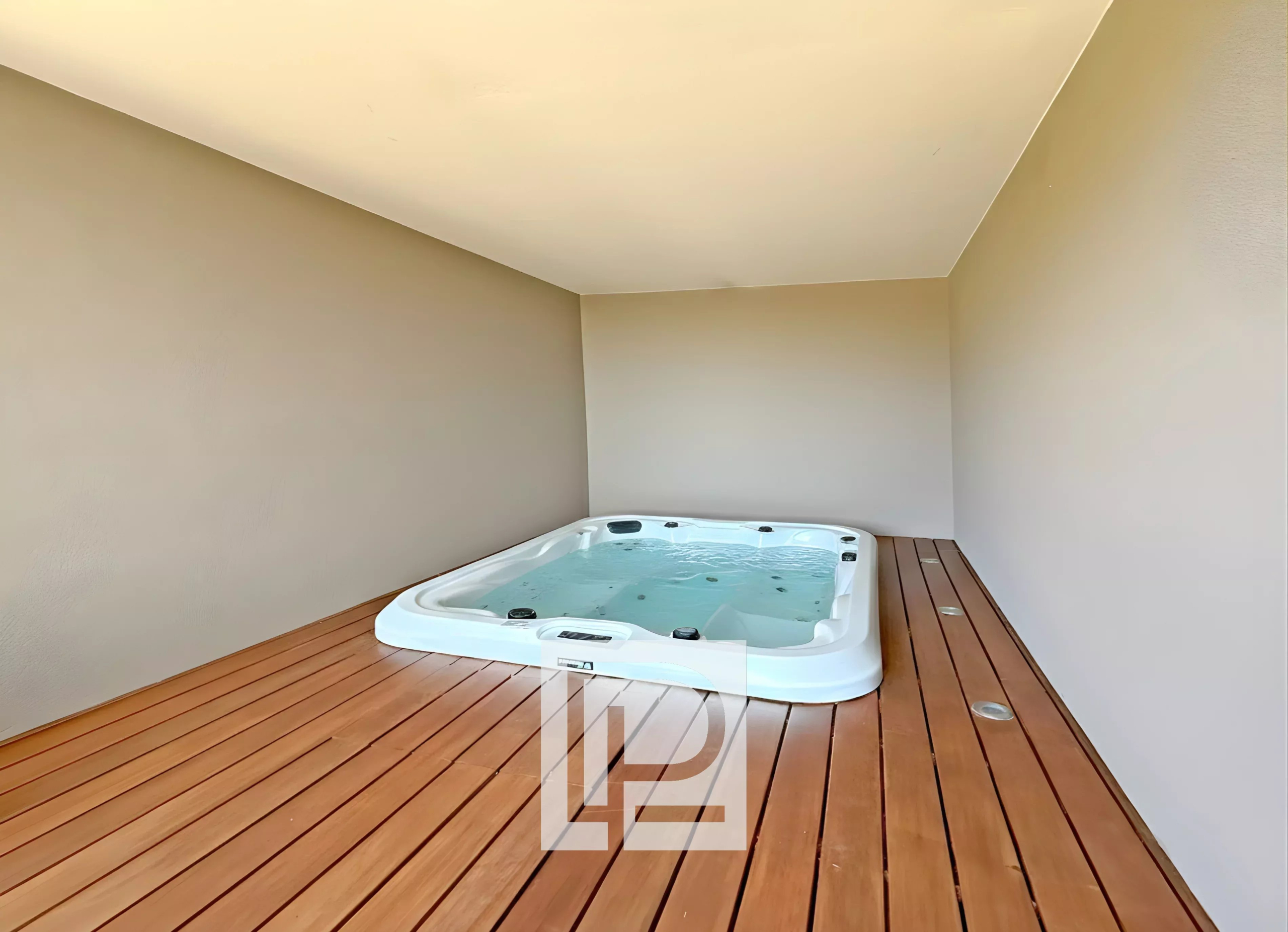 White rectangular hot tub filled with water sits on a dark wood deck inside a beige-walled, minimal outdoor patio or balcony.