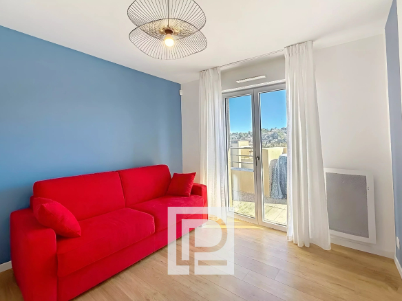 Bright living room with a red sofa against a blue wall, white curtains, and glass balcony doors letting in sunlight.