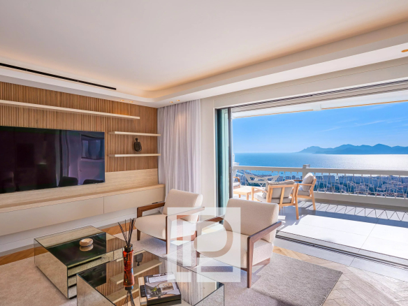 Modern living room with a wood-accent wall, large TV, beige seating, and glass coffee tables opening to an ocean-view balcony.