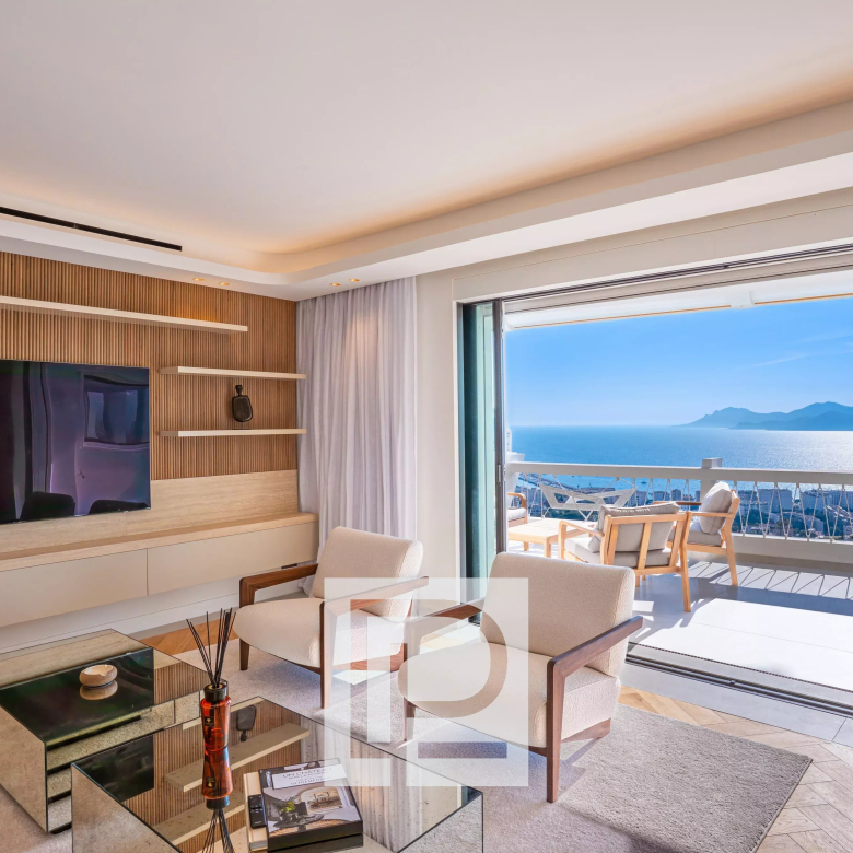 Modern living room with a wood-accent wall, large TV, beige seating, and glass coffee tables opening to an ocean-view balcony.