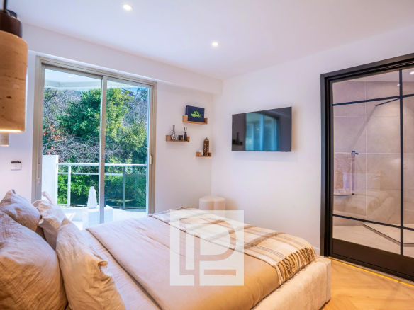 Modern bedroom with a beige bed, plush pillows, and a sliding glass door opening to a balcony with greenery outside, and a wall-mounted TV opposite the bed.