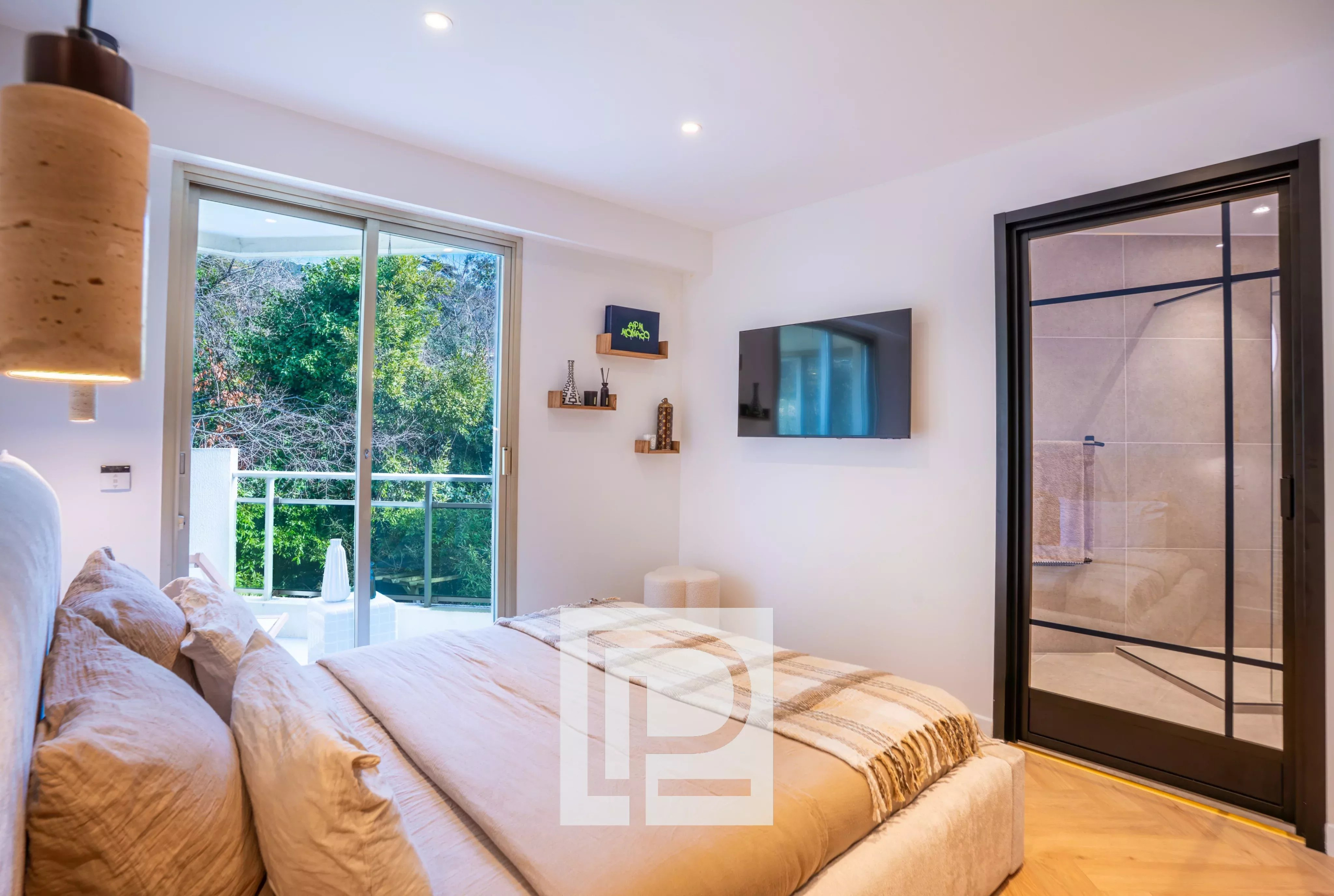 Modern bedroom with a beige bed, plush pillows, and a sliding glass door opening to a balcony with greenery outside, and a wall-mounted TV opposite the bed.
