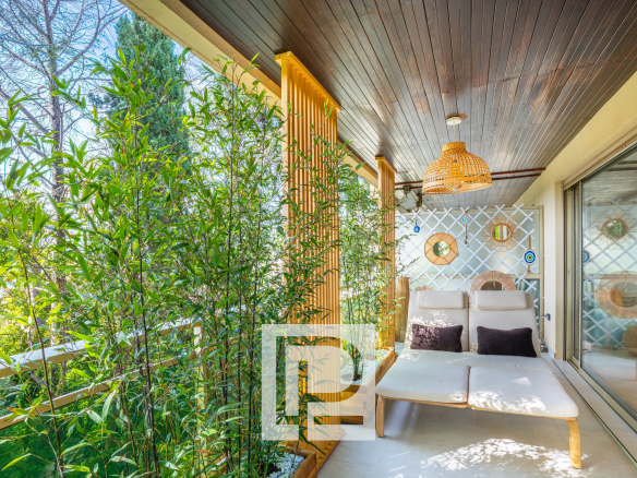 Balcony seating area with tall bamboo plants, a woven wicker pendant light, and cream lounge chairs with dark pillows under a wooden ceiling and sliding glass doors.