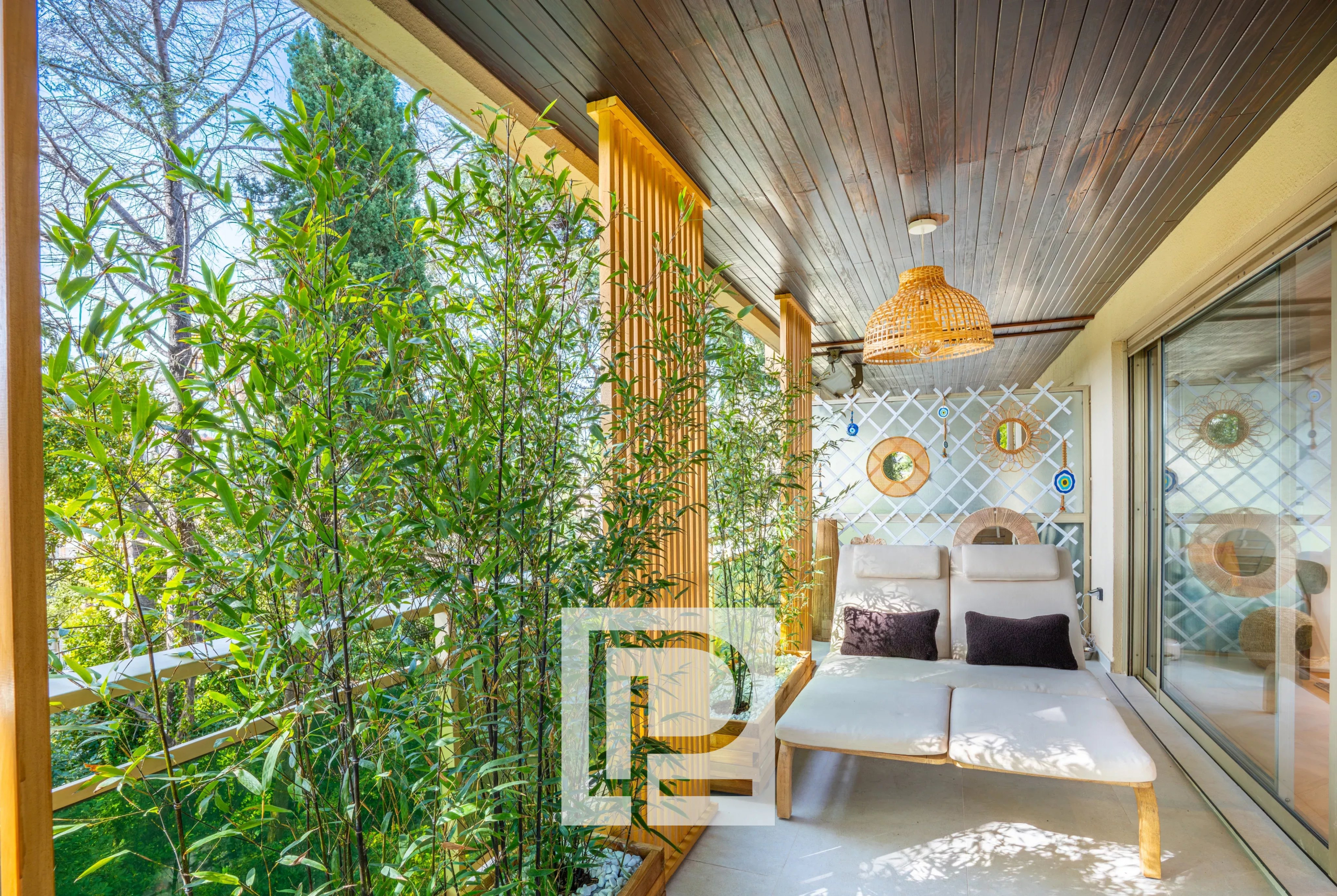 Balcony seating area with tall bamboo plants, a woven wicker pendant light, and cream lounge chairs with dark pillows under a wooden ceiling and sliding glass doors.