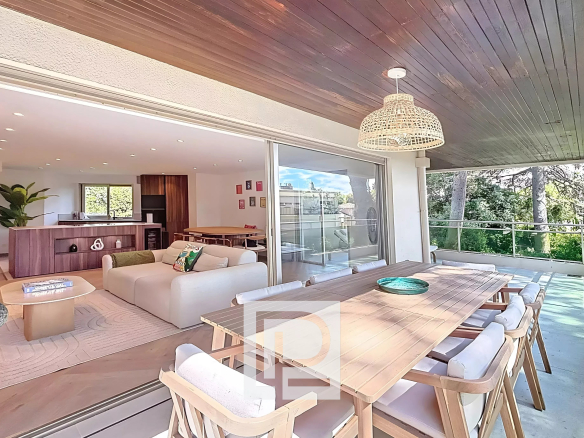 Open-plan living area connects to a covered patio: white sofa, wooden dining table, and kitchen island visible inside, with sliding doors between spaces.