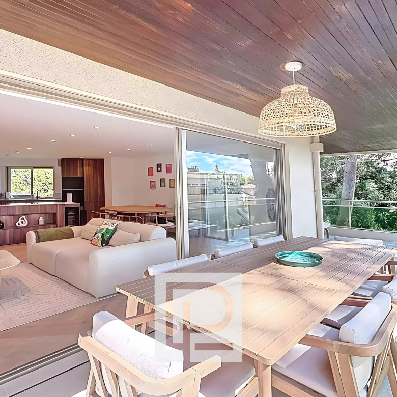 Open-plan living area connects to a covered patio: white sofa, wooden dining table, and kitchen island visible inside, with sliding doors between spaces.