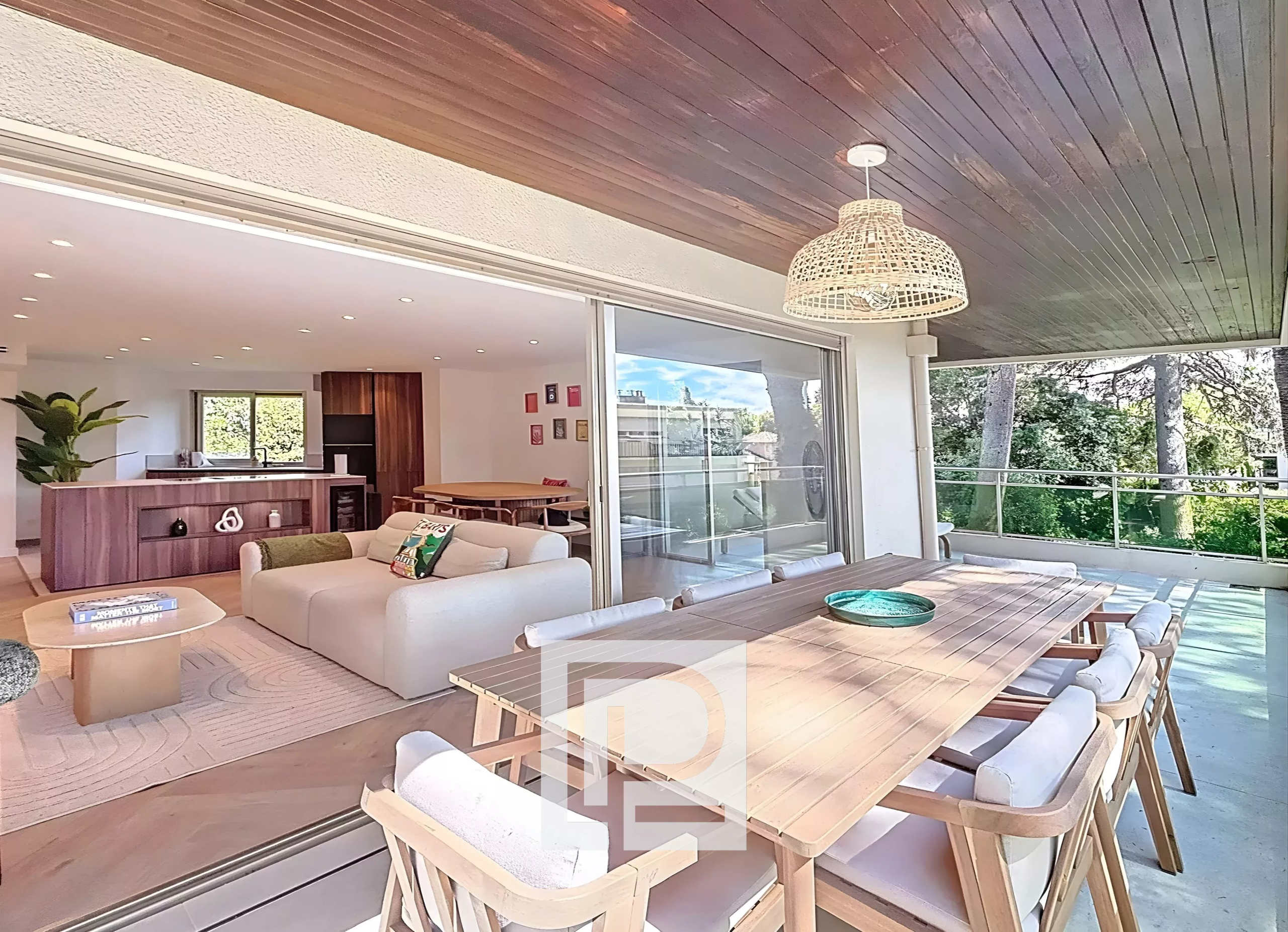 Open-plan living area connects to a covered patio: white sofa, wooden dining table, and kitchen island visible inside, with sliding doors between spaces.