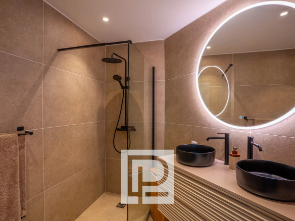 Modern bathroom with beige tiled walls, a glass shower enclosure, circular backlit mirror, and two black vessel sinks on a curved vanity.