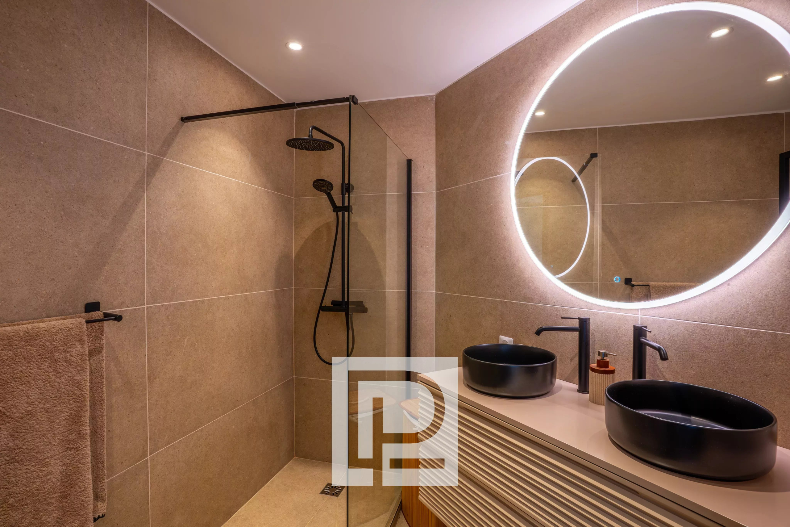 Modern bathroom with beige tiled walls, a glass shower enclosure, circular backlit mirror, and two black vessel sinks on a curved vanity.