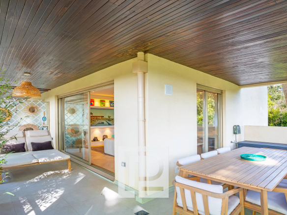 Outdoor patio with a dark wooden ceiling, beige seating, and a large wooden dining table on a sunny terrace.