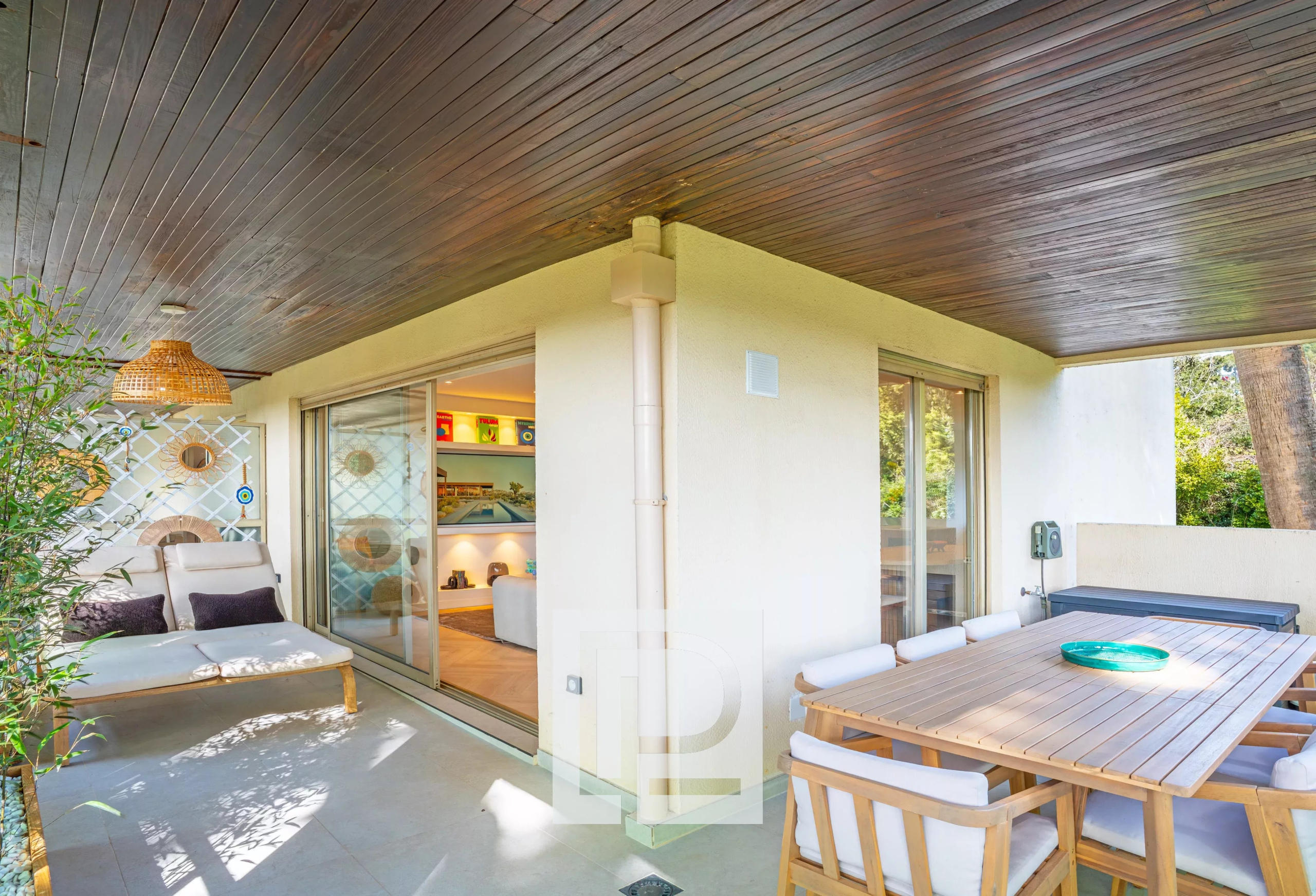 Outdoor patio with a dark wooden ceiling, beige seating, and a large wooden dining table on a sunny terrace.