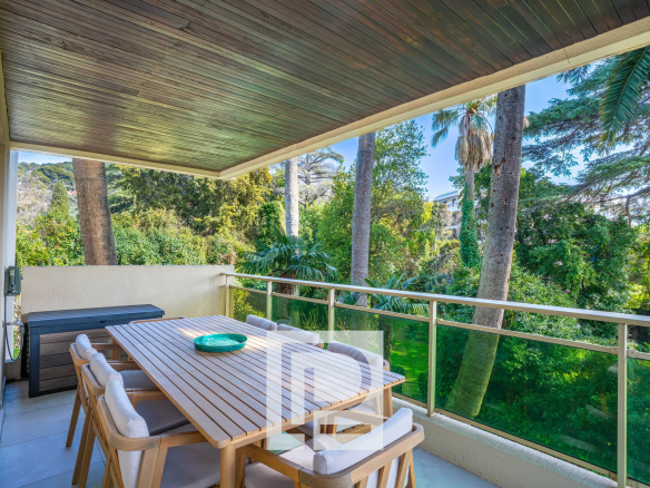 Outdoor balcony dining area with a long wooden table and cushioned chairs, overlooking a lush tropical garden with tall trees.