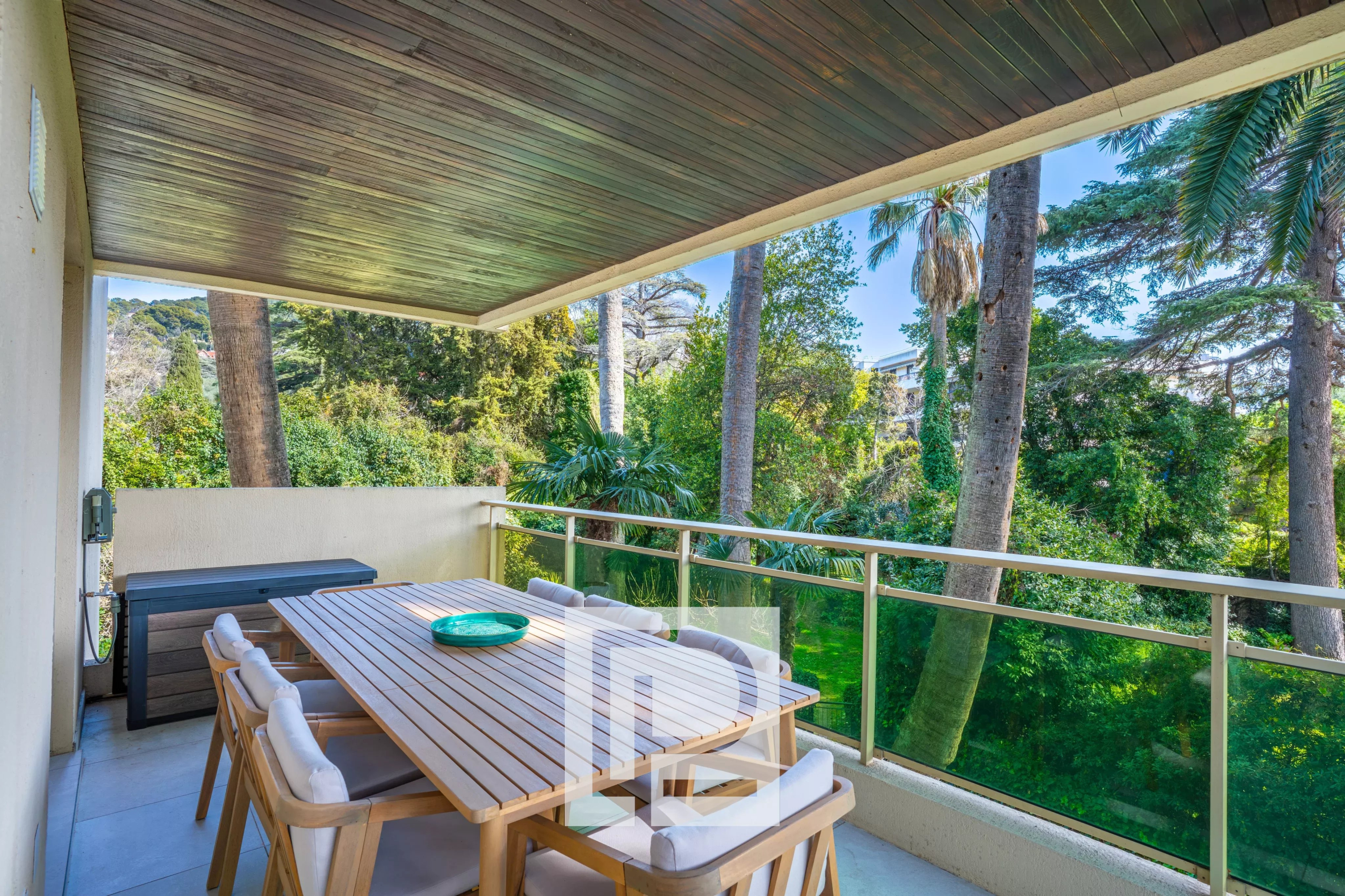 Outdoor balcony dining area with a long wooden table and cushioned chairs, overlooking a lush tropical garden with tall trees.