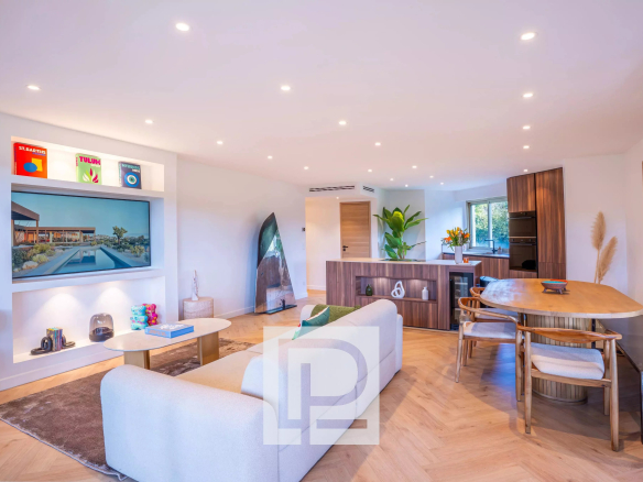 Open-plan living room with a white sofa, coffee table, and a wall-mounted TV, kitchen area in the background.