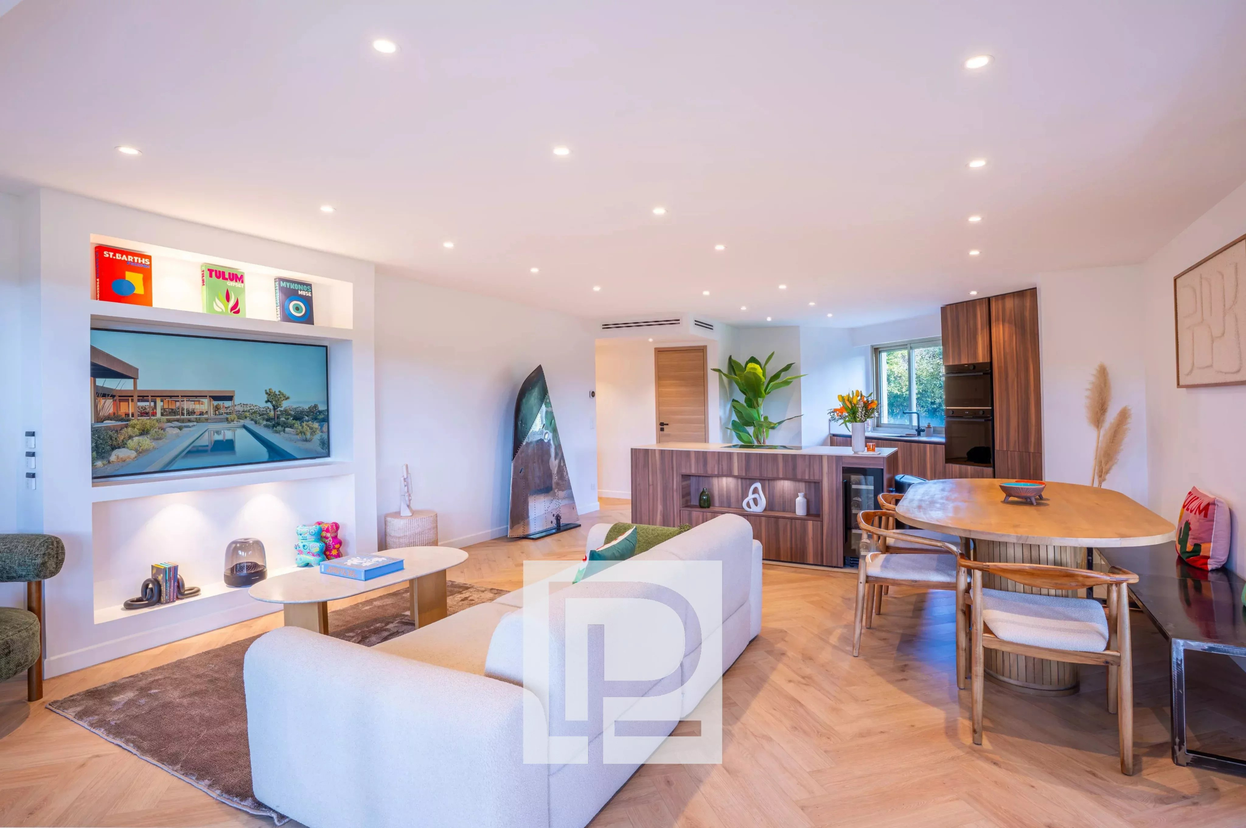 Open-plan living room with a white sofa, coffee table, and a wall-mounted TV, kitchen area in the background.