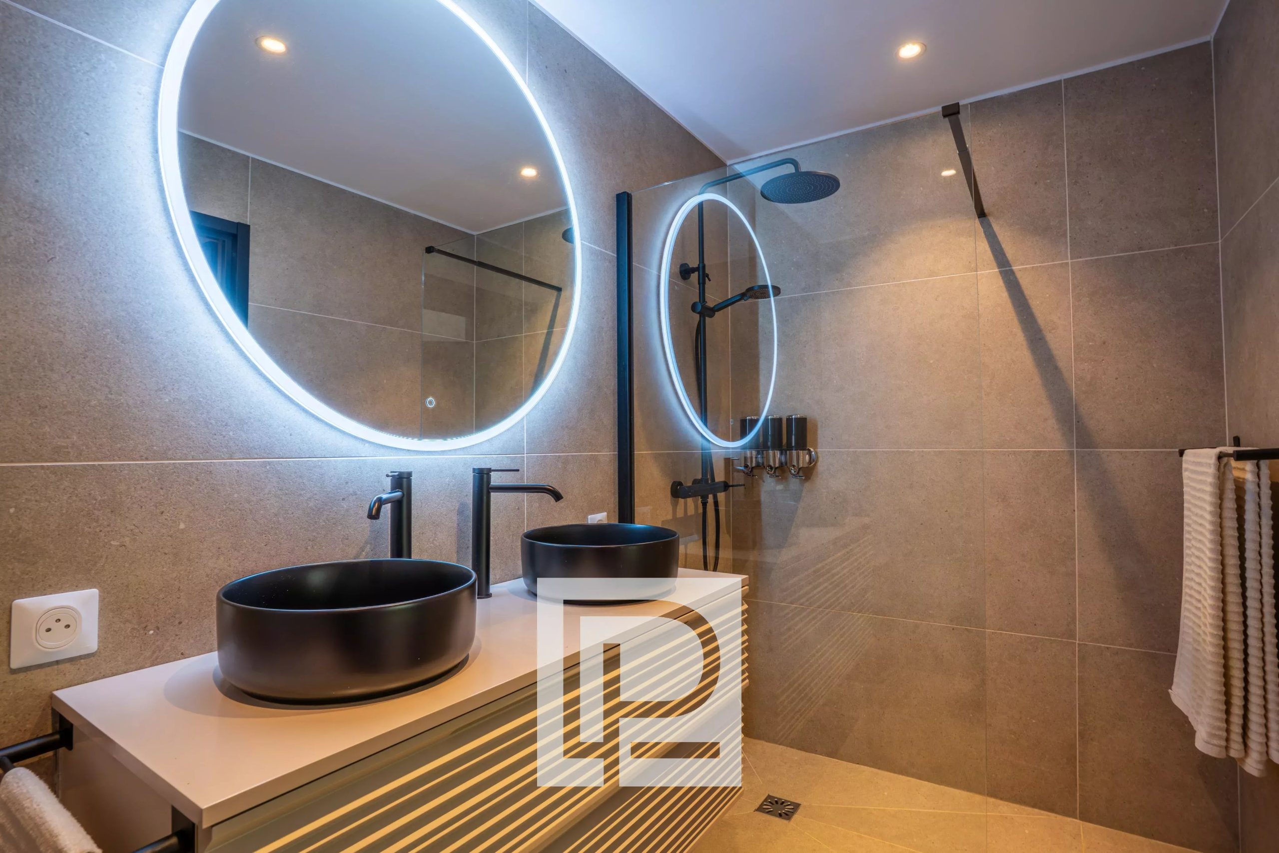 Modern bathroom with double black vessel sinks, backlit circular mirrors, and a glass shower.