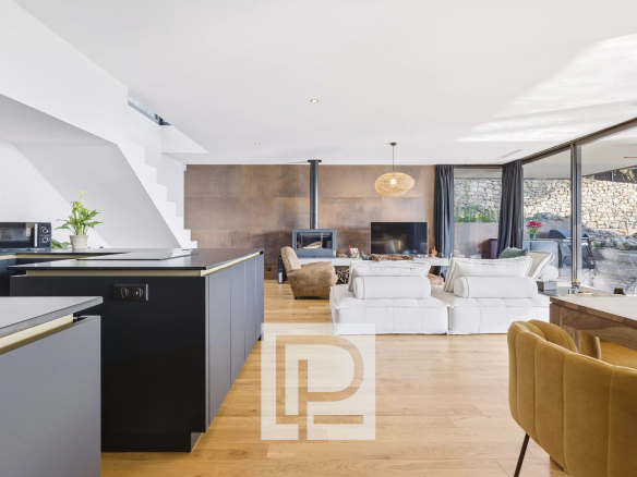 Spacious open-plan living area with a black kitchen island, white sofas, and a wood-burning stove against a copper-toned wall, opening to an outdoor patio via glass doors.