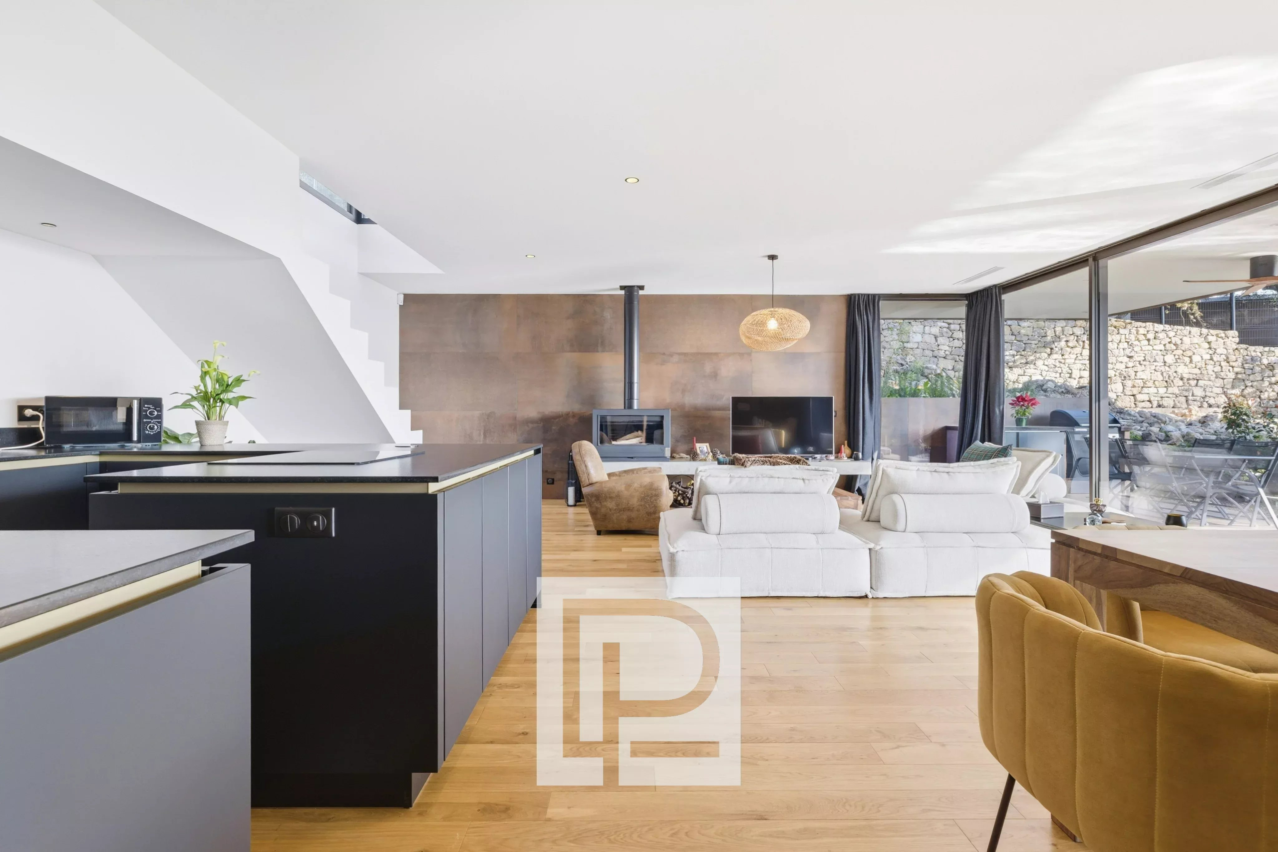 Spacious open-plan living area with a black kitchen island, white sofas, and a wood-burning stove against a copper-toned wall, opening to an outdoor patio via glass doors.