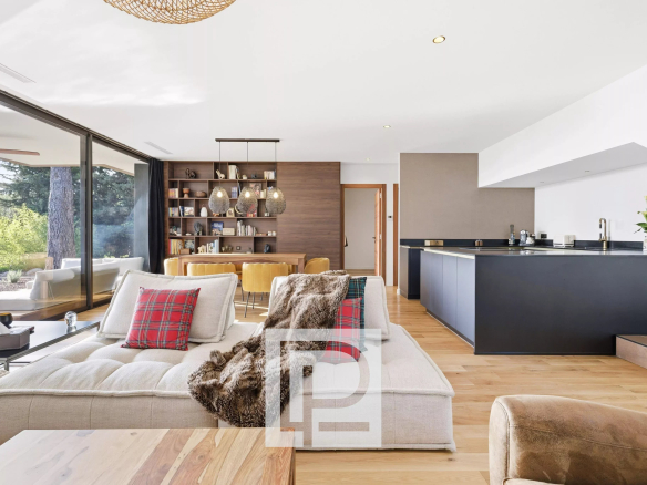 Open-plan living room with a beige sectional, plaid red cushions, fur throw, and a dark kitchen island; large glass doors open to greenery outside.
