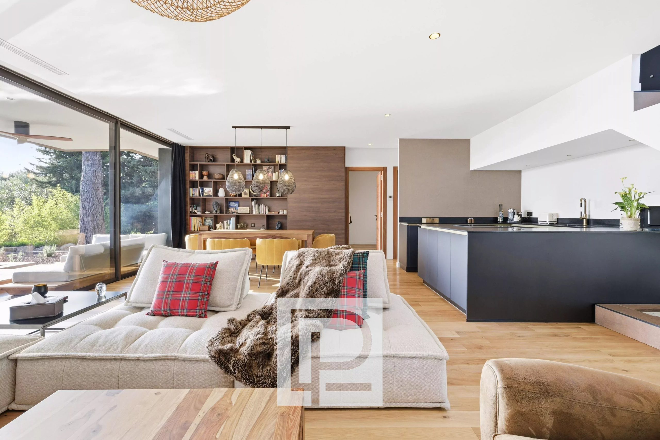 Open-plan living room with a beige sectional, plaid red cushions, fur throw, and a dark kitchen island; large glass doors open to greenery outside.