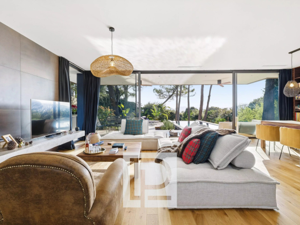 Bright, open-concept living room with large glass doors opening to a sunny outdoor area, featuring a light gray sectional with plaid cushions and a brown leather chair.