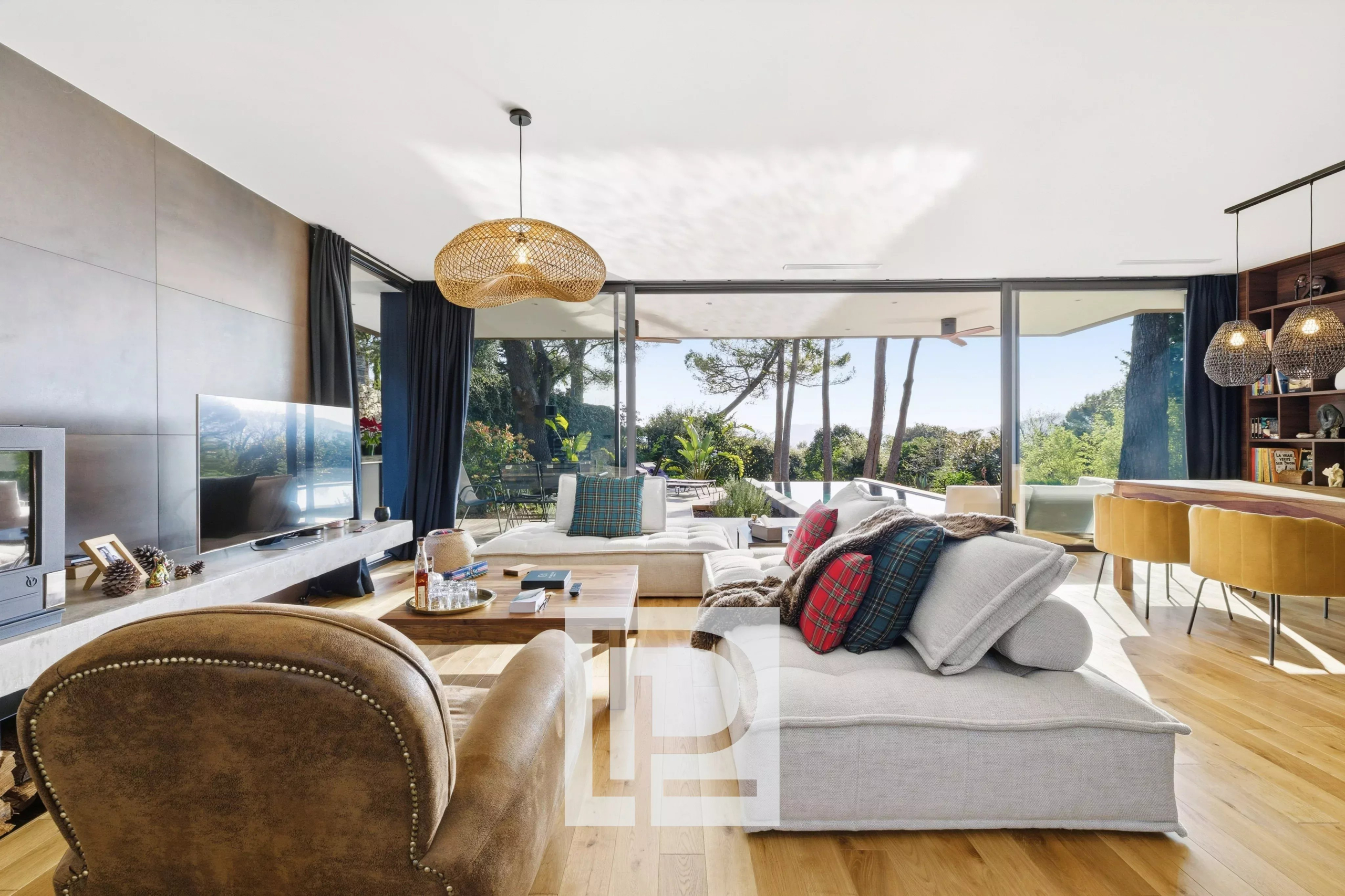 Bright, open-concept living room with large glass doors opening to a sunny outdoor area, featuring a light gray sectional with plaid cushions and a brown leather chair.