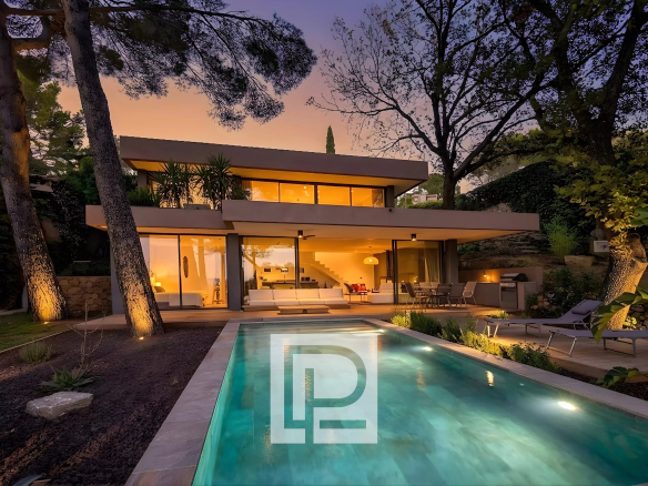 Modern two-story house with large glass walls, warmly lit interior, surrounded by trees at dusk, pool in foreground and outdoor seating area on the patio.