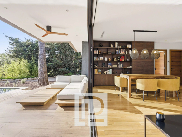 Open-plan living-dining area split by a glass wall: outdoor white sectional, wooden deck, and pool beside an interior with a large bookshelf and mustard chairs around a wooden table.