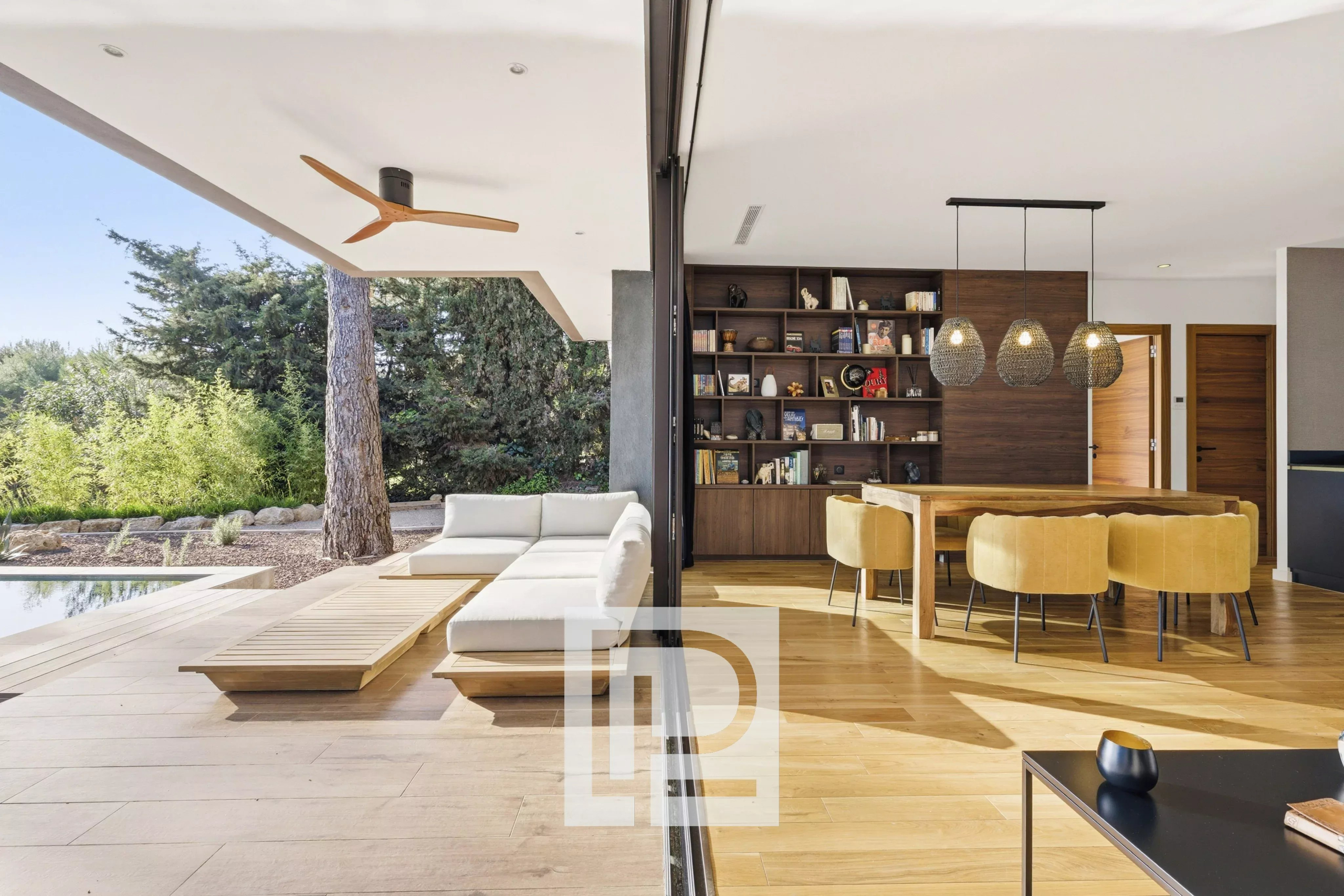 Open-plan living-dining area split by a glass wall: outdoor white sectional, wooden deck, and pool beside an interior with a large bookshelf and mustard chairs around a wooden table.