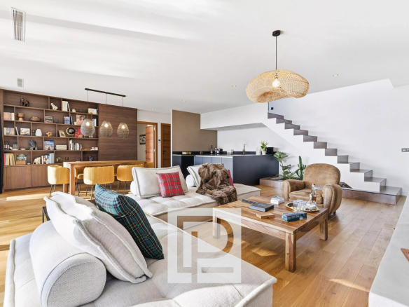 Open-plan living area with a beige sofa, plaid pillows, wooden coffee table, and a large bookshelf wall next to a dining table with mustard chairs under pendant lights on a light wooden floor.