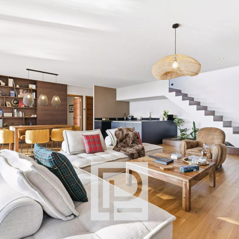 Open-plan living area with a beige sofa, plaid pillows, wooden coffee table, and a large bookshelf wall next to a dining table with mustard chairs under pendant lights on a light wooden floor.
