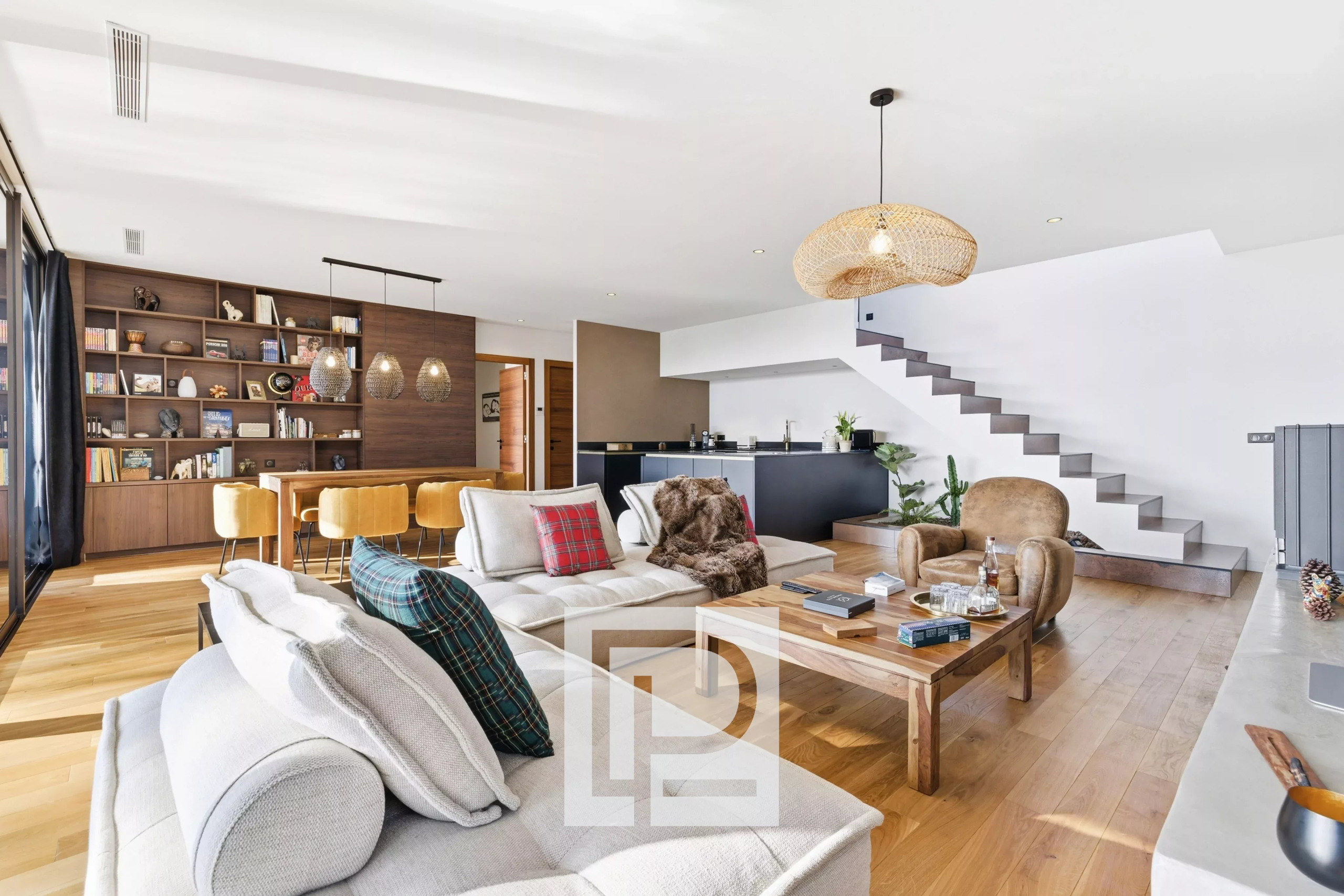 Open-plan living area with a beige sofa, plaid pillows, wooden coffee table, and a large bookshelf wall next to a dining table with mustard chairs under pendant lights on a light wooden floor.