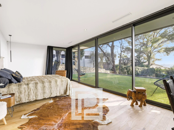 Bright bedroom with floor-to-ceiling glass doors overlooking a green backyard; bed with a fur throw and dark pillows, plus a cowhide rug on light wood floors.