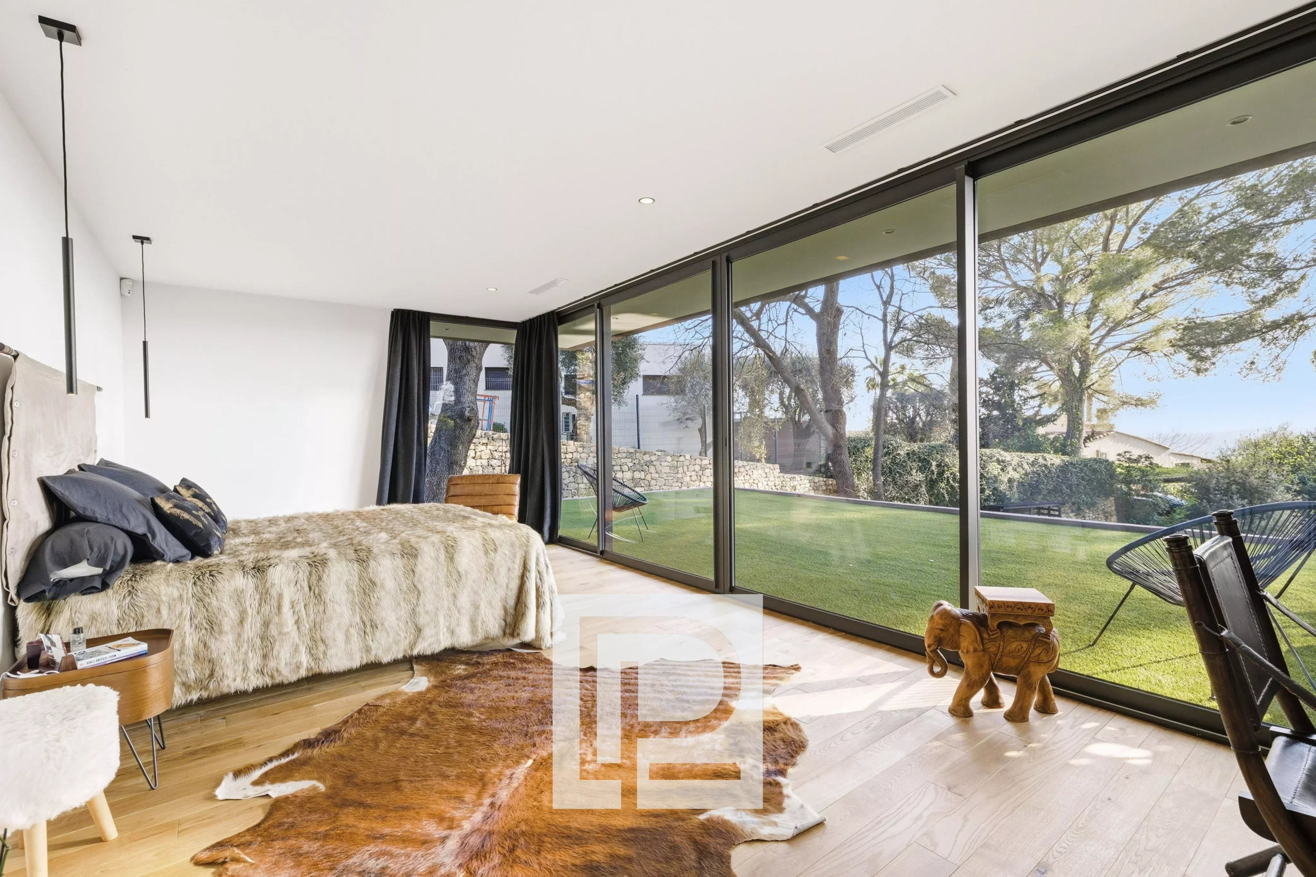 Bright bedroom with floor-to-ceiling glass doors overlooking a green backyard; bed with a fur throw and dark pillows, plus a cowhide rug on light wood floors.