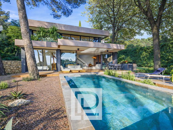 Modern two-story house with glass walls, open-plan living area, and a rectangular in-ground pool surrounded by a gravel yard and trees.