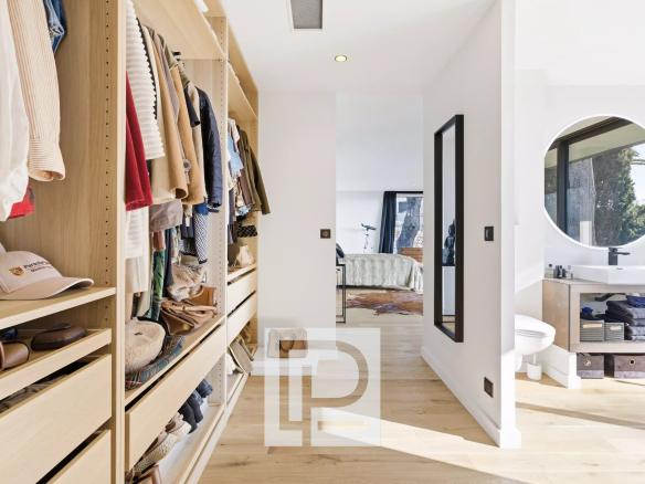 Walk-in closet with light wood shelves and hanging clothes, leading to a bright bedroom at the end of the hallway.