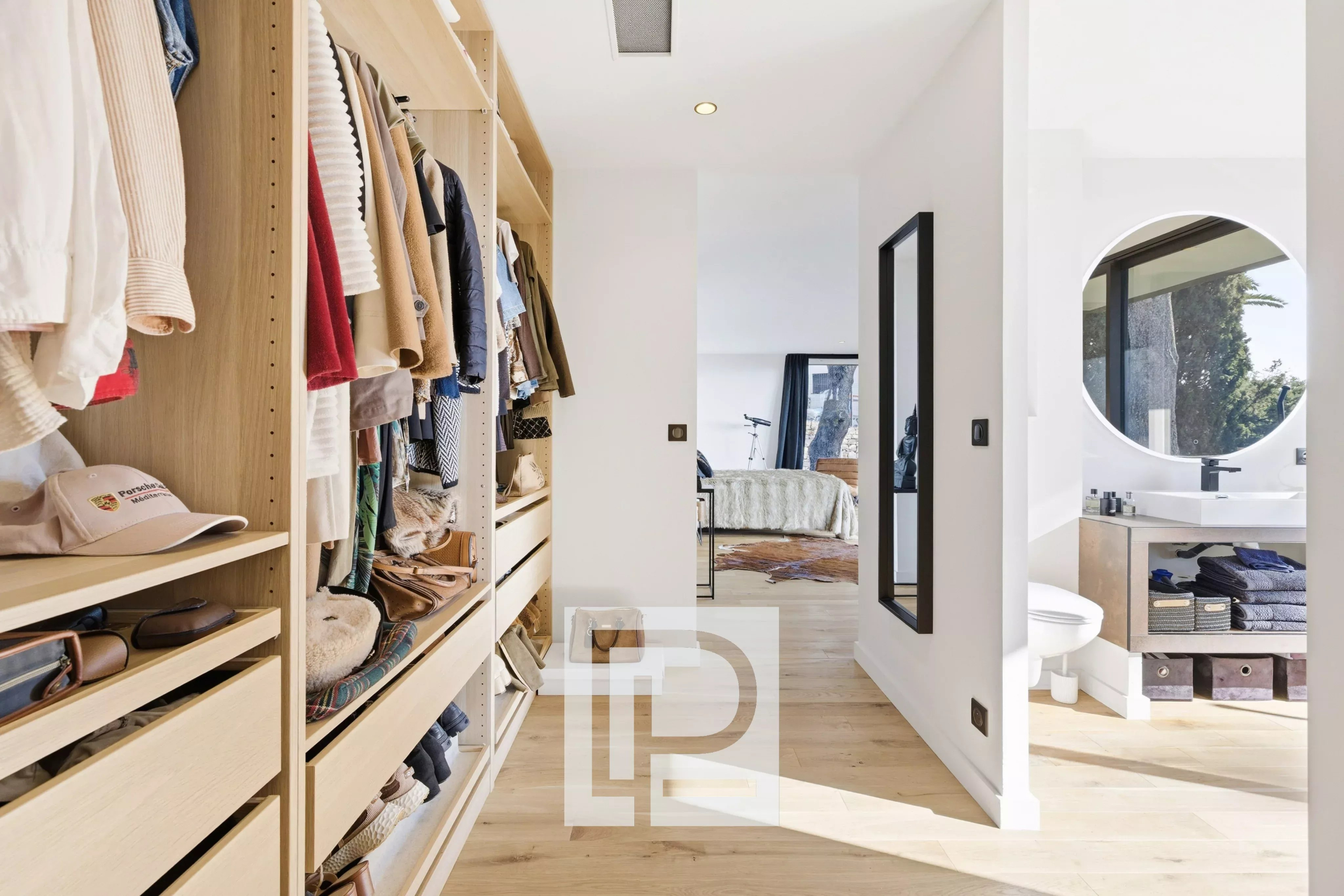 Walk-in closet with light wood shelves and hanging clothes, leading to a bright bedroom at the end of the hallway.