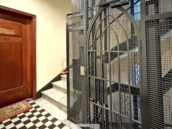 Narrow building hallway with a tall metal cage elevator gate, checkered black-and-white floor tiles, a wooden door on the left, and marble stairs behind the gate.
