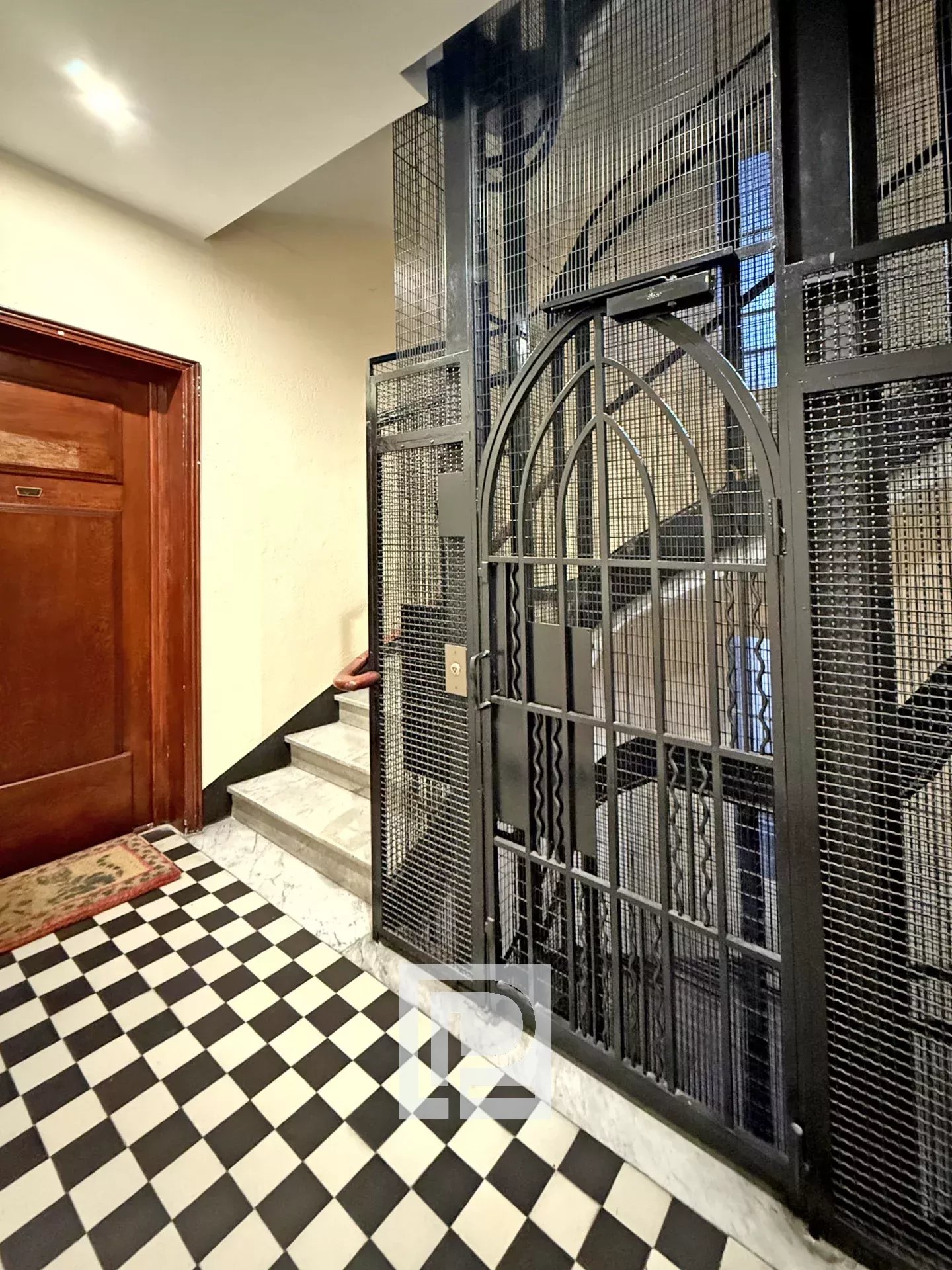 Narrow building hallway with a tall metal cage elevator gate, checkered black-and-white floor tiles, a wooden door on the left, and marble stairs behind the gate.